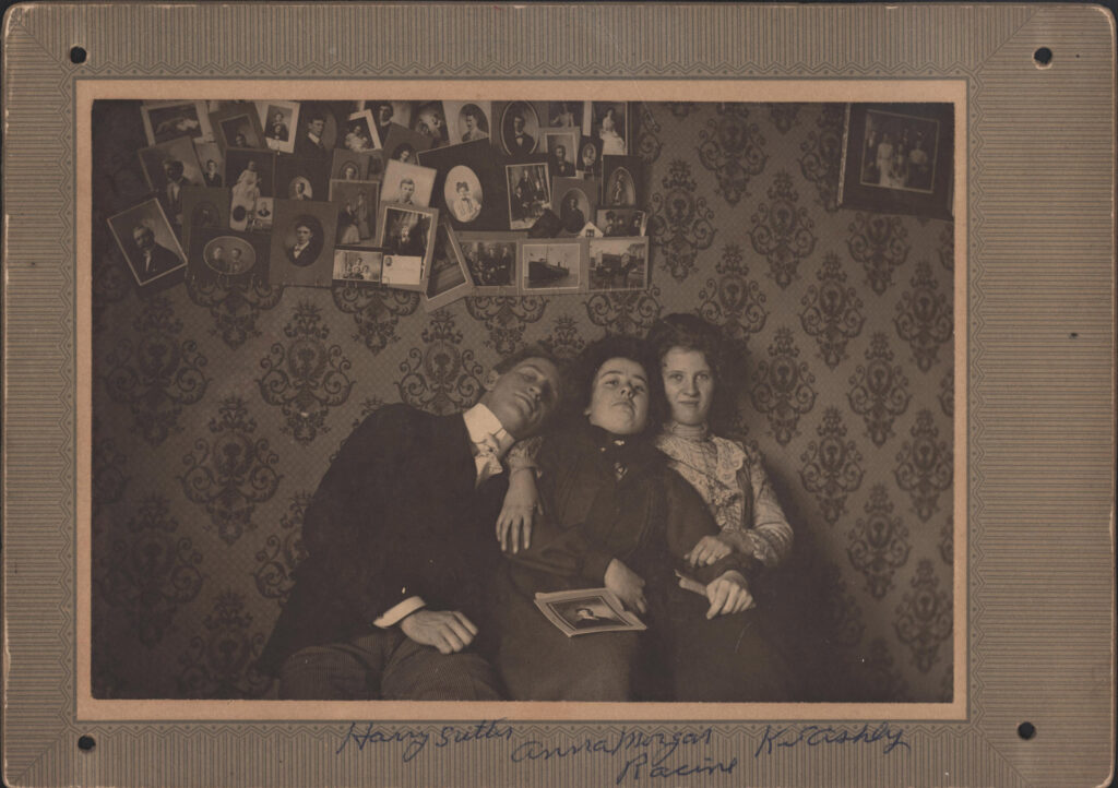 Three young people are posing at Christmas, surrounded by family photos on a wall, and one photo being held by the young woman in the middle.  The woman on the right appears to have blond hair and light-colored eyes. She also seems to be wearing a wedding ring. The woman in the middle is not wearing a wedding ring. 
The names handwritten along the bottom appear to be: Harry Sutler (or Sutter), Anna Morgan, and K S Ashly. Underneath these names is the word Racine.
