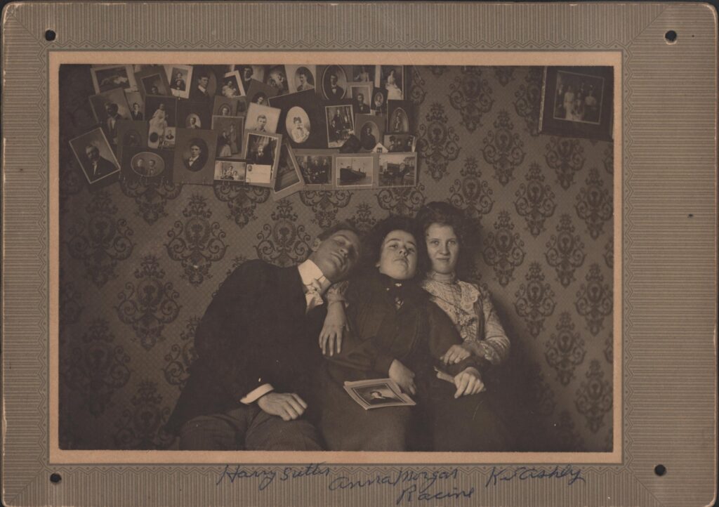 Three young friends, with Anna Morgan in the center, pose for a photograph against a wall with fancy wallpaper and 30 plus photos pinned on the wall.